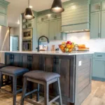 A newly painted kitchen with sage green cabinets, bright lights, and hardwood floors