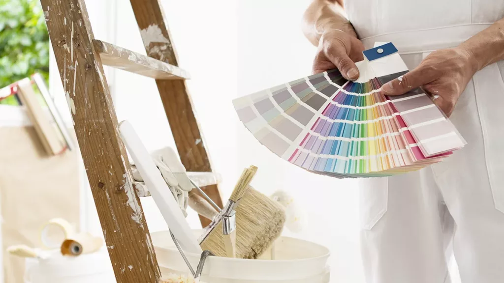 A professional residential painter dressed in white stands next to a ladder, holding a spread of paint color palette samples, showing the concept of color psychology